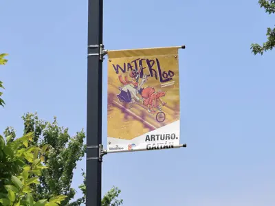 A street banner with colourful artwork hangs from a pole on a sunny day, surrounded by trees.