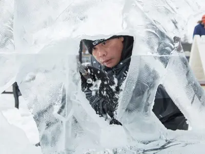 An artist carves a large ice sculpture outdoors in the snow.